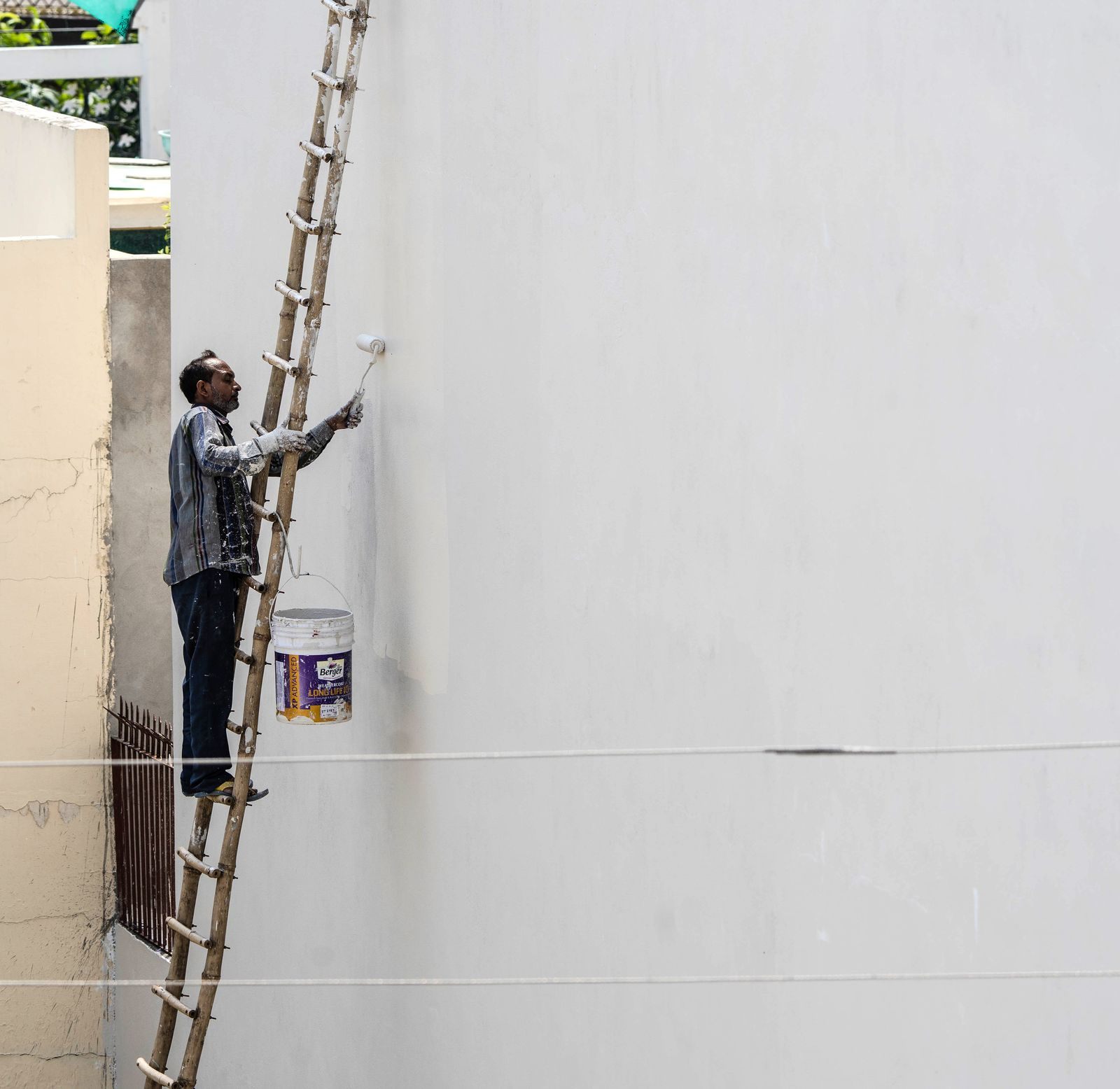 A man on a ladder painting a building's exterior wall with a roller brush outdoors